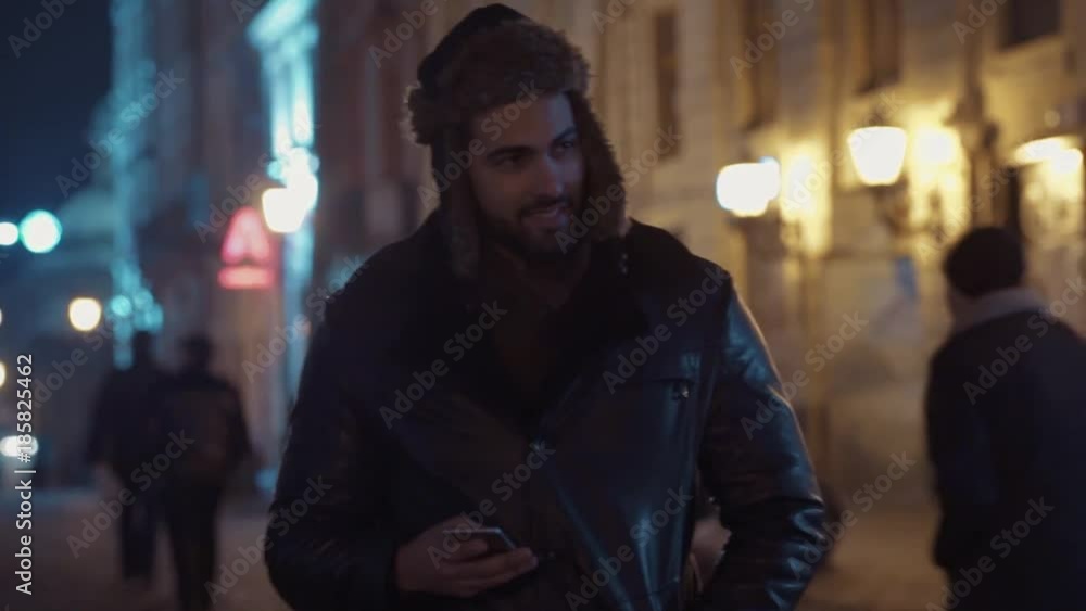 Good-looking bearded young man walking down the street, using his smartphone and smiling towards the screen. City lights on the background. Holidays, modern technologies Good-looking bearded young man walking down the street, using his smartphone and smiling towards the screen. City lights on the background. Holidays, modern technologies