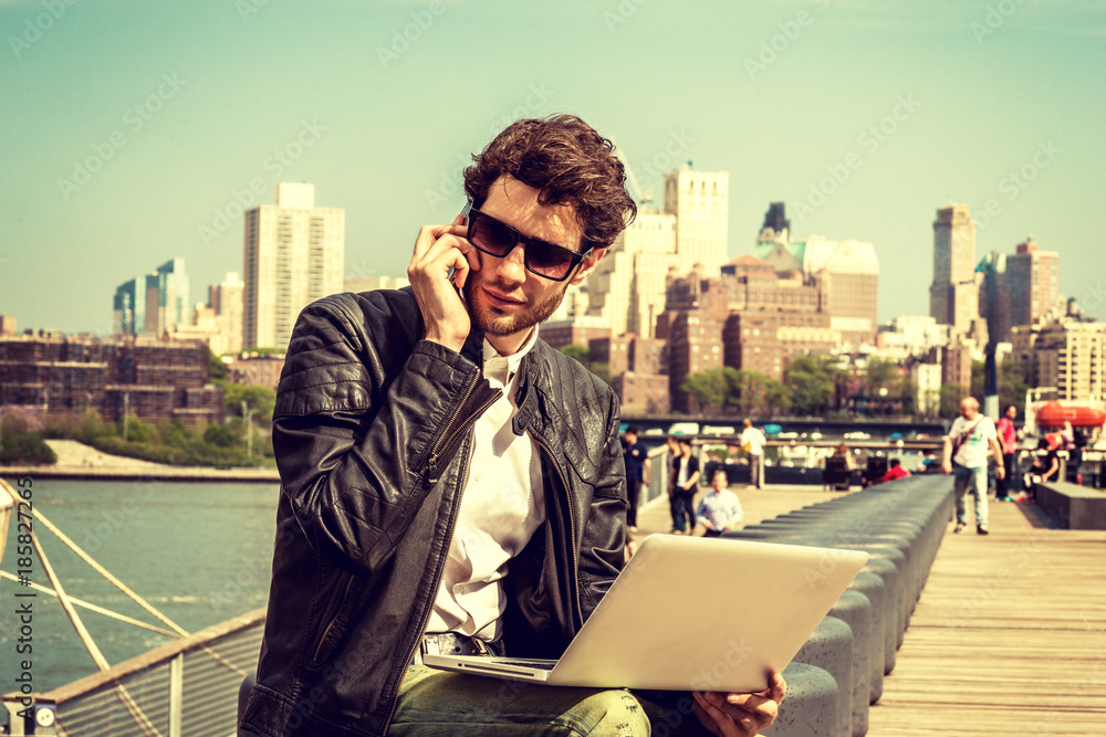 Businessman traveling, working in New York. Wearing sunglasses, a young guy with beard, sitting on bench at harbor, working on laptop computer, talking on phone in same time. Retro filtered look.. Businessman traveling, working in New York. Wearing sunglasses, a young guy with beard, sitting on bench at harbor, working on laptop computer, talking on phone in same time. Retro filtered look..