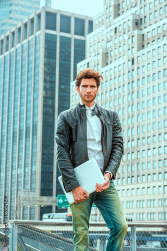 European businessman traveling, working in New York. Wearing black leather jacket, blue jeans, hand carrying laptop computer, a young guy with beard, standing in business district with high buildings © Alexander Image