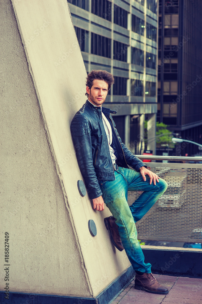 Man Urban Casual Fashion. Wearing black leather jacket, blue jeans, brown  boot shoes, a young guy with beard, leaning against wall on balcony, facing  street, thinking. City Boy. Filtered effect.. Stock Photo