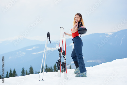 Attractive lady skier wearing ski pants and red bodice, smiling to the camera, standing on the top of the snowy slope with ski equipment at winter ski resort in the mountains