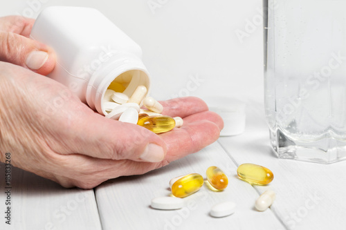 Woman hands hold vitamin complex and dietary supplement, pours from white bottle into palm yellow capsules of omega 3, white pills of calcium and glucosamine, glass of water on wooden planks table