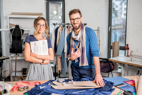 Portrait of a couple of fashion designers working with fabric and clothing sketches at the studio full of tailoring tools and equipment