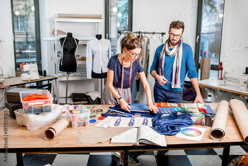 Couple of fashion designers working with fabric and clothing sketches at the studio full of tailoring tools and equipment
