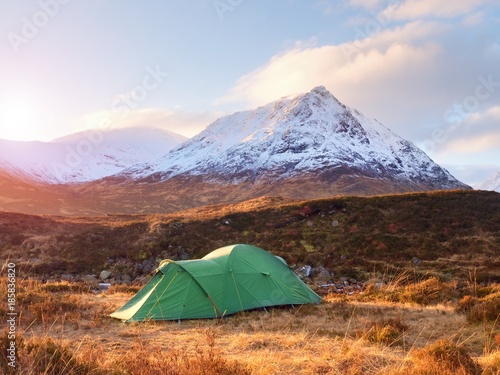 Green touristic tent on meadow at river below snowy cone of mountain Stob Dearg 1021 metres high