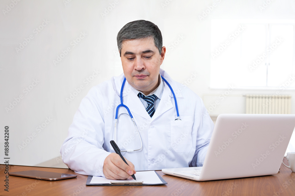 Handsome male doctor works in the office at a computer and writes in a workbook Handsome male doctor works in the office at a computer and writes in a workbook