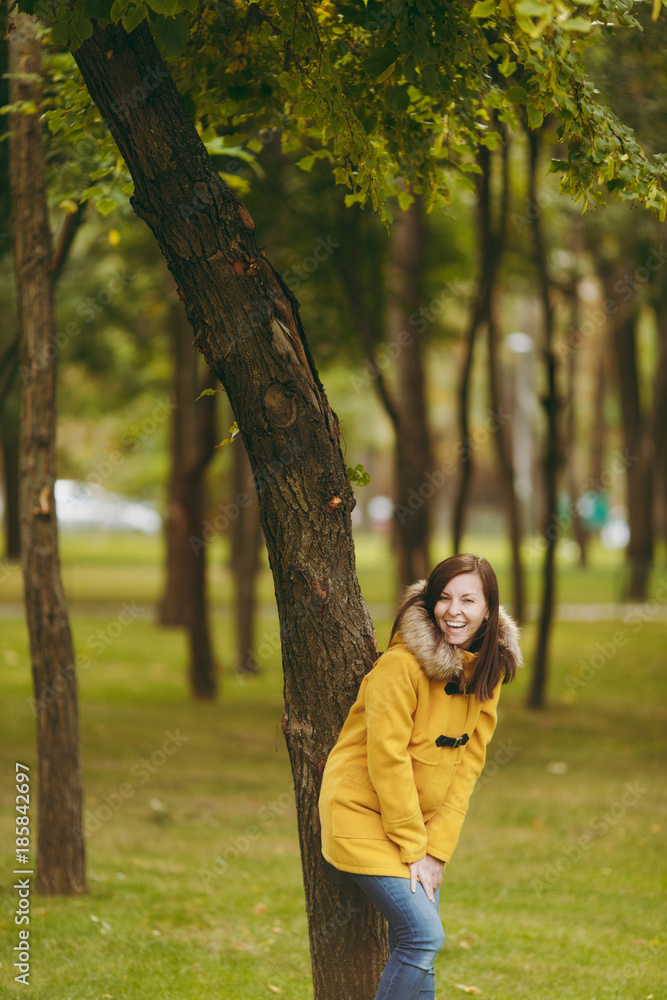 Beautiful happy caucasian young smiling brown-hair woman in yellow coat, jeans, boots in green forest. Fashion female model with fall golden leaves standing and walking in early autumn park outdoors.