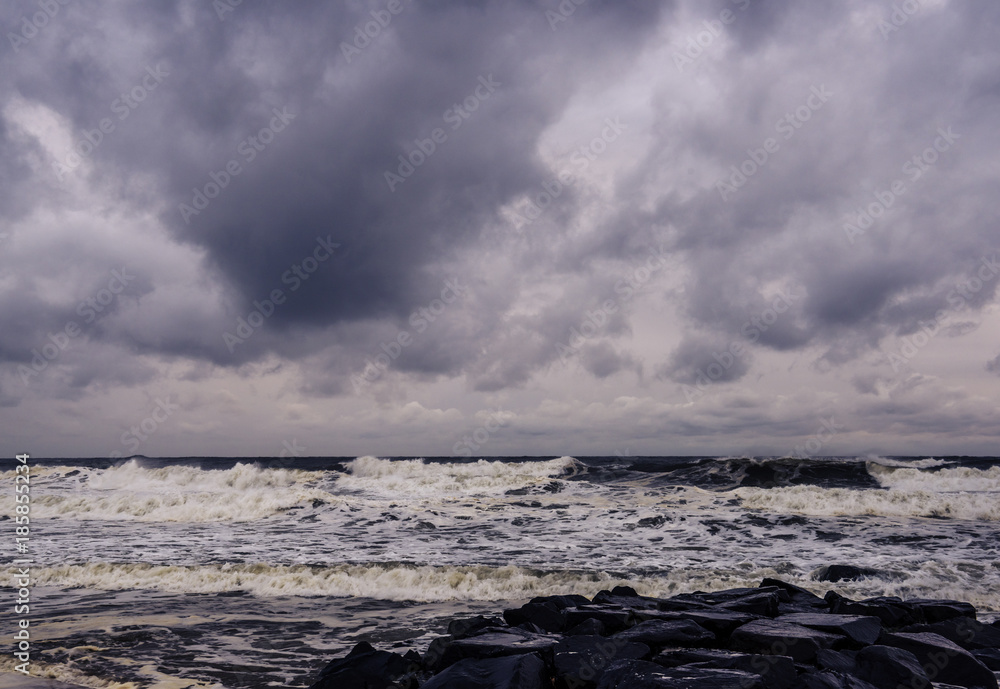Moody morning on Manasquan beach at the end of the summer season. Ocean waves and stones on the foreground and cloudy sky on the background. Dark emotional colors.