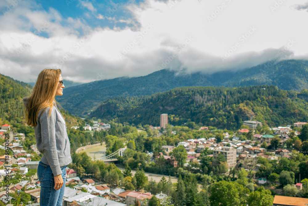 Naklejka premium So a beautiful woman is standing on top of a mountain in a hoodie. looking at the mountains and trees from above.Beautiful panorama. Borjomi. Far away city. On top of the mountains and clouds