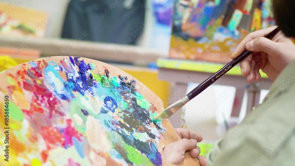A close-up of the artist's hands holds a brush and a palette. Draw in paints. In the studio. Canvas. A close-up of the artist's hands holds a brush and a palette. Draw in paints. In the studio. Canvas.