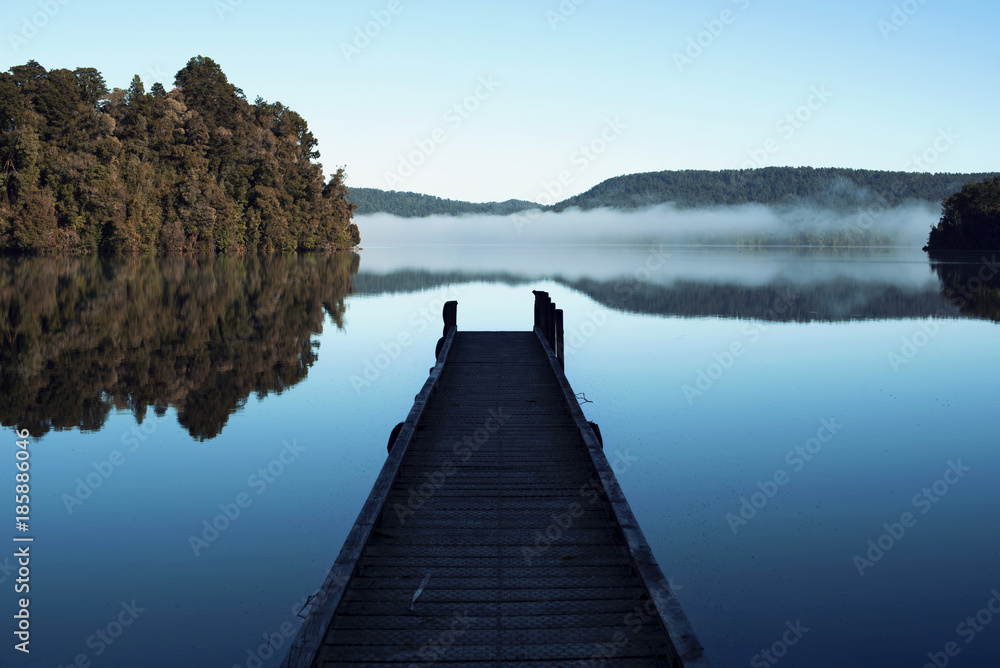 Lago en calma con pequeño embarcadero y reflejo de niebla y árboles.