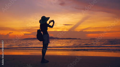 Silhouette of young tourist woman in hat taking photo with cellphone during sunset in ocean beach