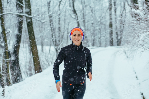 Running sport woman enjoying winter. Female runner jogging in cold winter forest wearing black sporty running clothing and listening music with headphones. Snowfall