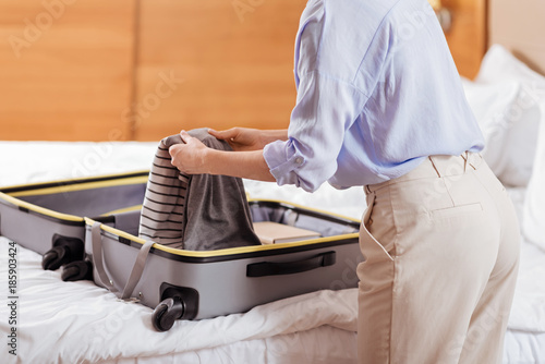 Clothes in luggage. Woman putting her elegant modern comfort clothes while standing near the bed when baggage lying on it 