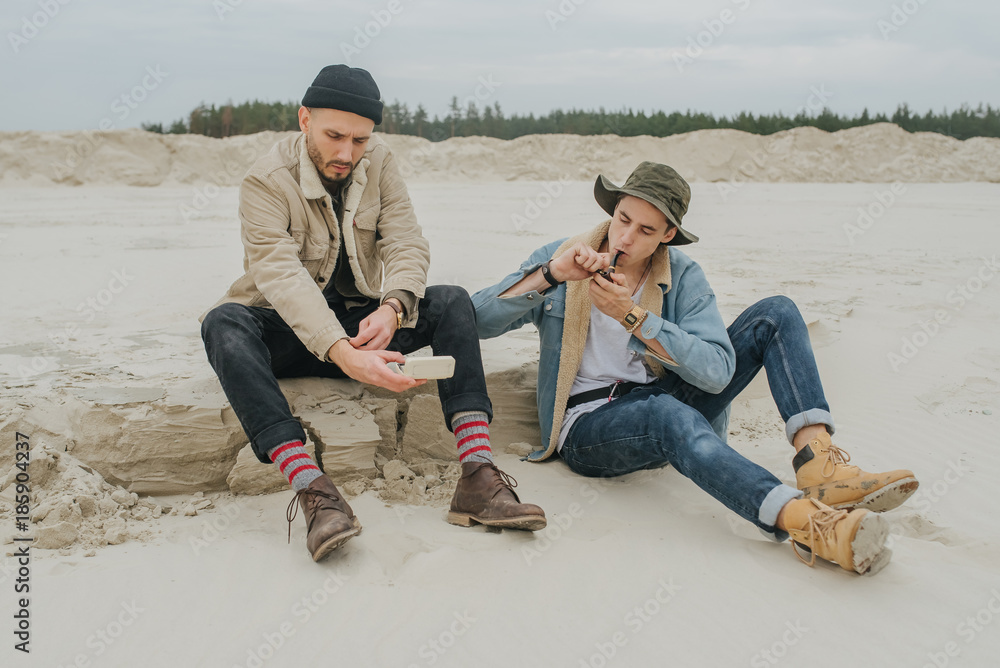 Hipsters sits on a land in the forest and playing the harmonica. Man and harmonica. A smoke break with a tourist. Rest after a hard road. Two travellers.