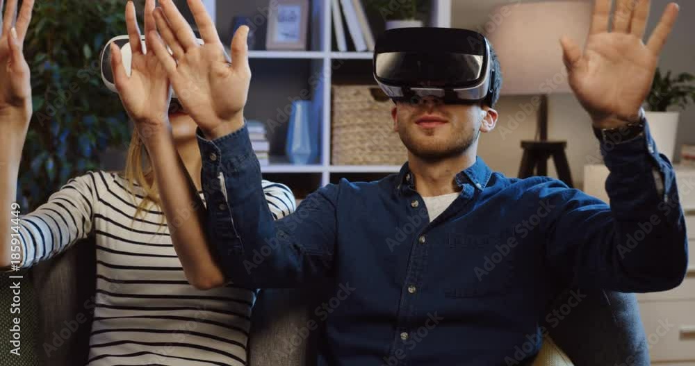 Young couple sitting on the couch in the living room in VR glasses and having VR headset, scrolling and taping in the air. Evening time. Close up. Portrait. Inside Young couple sitting on the couch in the living room in VR glasses and having VR headset, scrolling and taping in the air. Evening time. Close up. Portrait. Inside