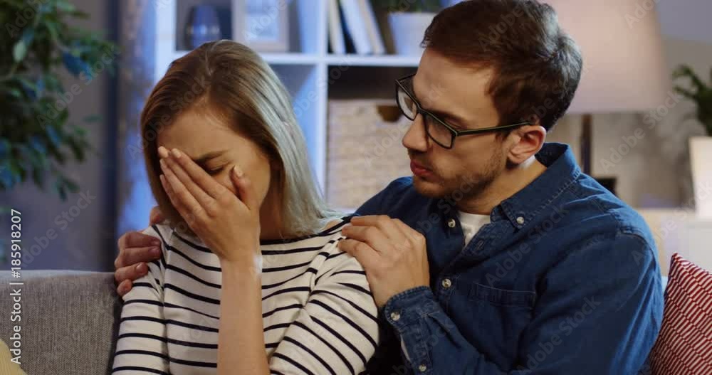 Portrait shot of the caucasian man in glasses hugging the blonde crying woman while they sitting on the sofa in the home room at night. Close up. Indoor Portrait shot of the caucasian man in glasses hugging the blonde crying woman while they sitting on the sofa in the home room at night. Close up. Indoor
