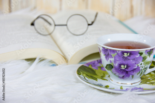 hot tea in vintage pansy floral pattern tea cup on saucer and white fur with old-fashioned glasses on open book