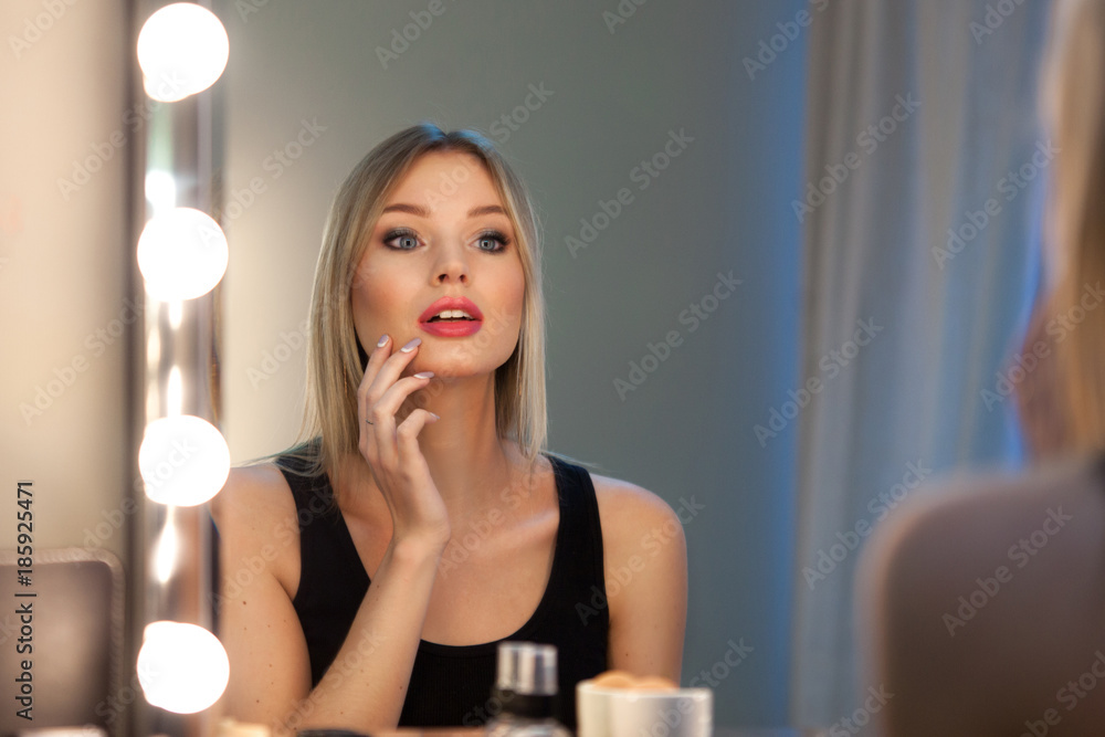 Attractive blonde girl primp at the mirror with lamps in the beauty studio. A happy satisfied customer of make-up and hairstyle service. Beautiful face with natural makeup and straight hair. Attractive blonde girl primp at the mirror with lamps in the beauty studio. A happy satisfied customer of make-up and hairstyle service. Beautiful face with natural makeup and straight hair.