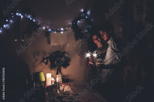 happy new year and merry christmas concept. happy stylish hipster couple holding burning sparklers fireworks and embracing in room at hoome in evening. happy holidays. toned