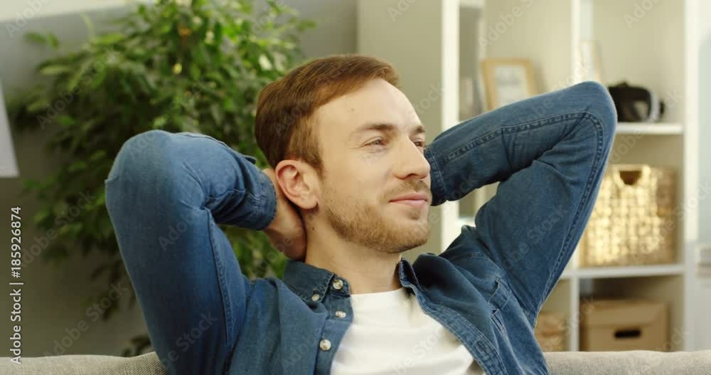 Close up of the handsome caucasian man in jeans shirt putting his hands over the head and having a rest at home. Portrait shot. Indoors Close up of the handsome caucasian man in jeans shirt putting his hands over the head and having a rest at home. Portrait shot. Indoors