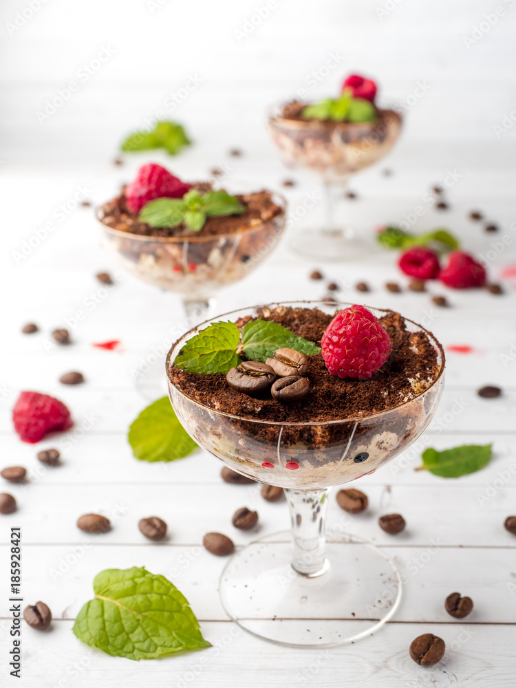 Dessert Tiramisu in glass goblet with mint coffee beans and fresh raspberries on white wooden table Dessert Tiramisu in glass goblet with mint coffee beans and fresh raspberries on white wooden table