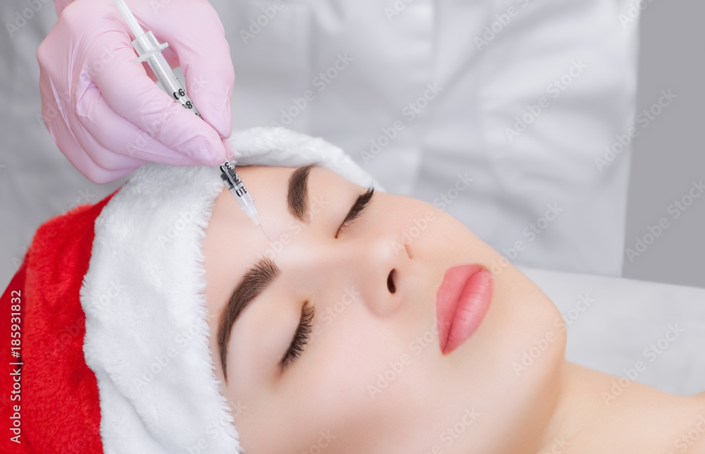Naklejka premium The cosmetologist makes the Botulinum toxin injection on the face skin of a beautiful, young woman in the Santa Claus hat. New Year's and Cosmetology concept