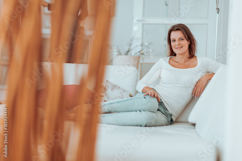 young woman in her late twenties sitting and laying on a lether white sofa in a cosy interier of her bright home and drinking a cup of tea and reading a book  totaly relaxed