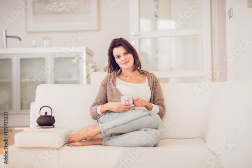 young woman in her late twenties sitting and laying on a lether white sofa in a cosy interier of her bright home and drinking a cup of tea and reading a book, totaly relaxed
