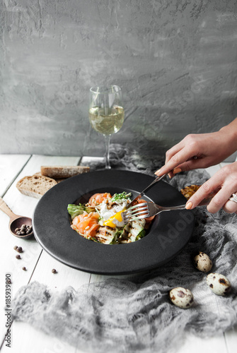 Salad with fresh salmon, grilled zucchini and hand that cuts the poached egg on a black plate on a concrete gray background