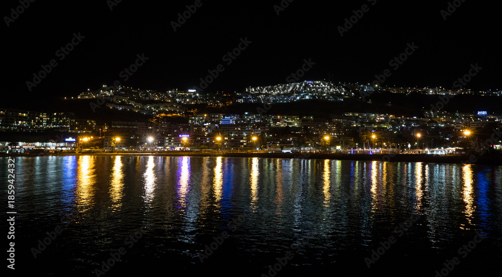 Night photography of the city with blue and yellow lights reflecting in the ocean. Puerto Rico, Gran Canaria, Spain.