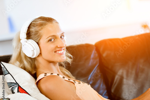 Young smiling woman with headphones relaxing on the sofa listening to music online using a smartphone