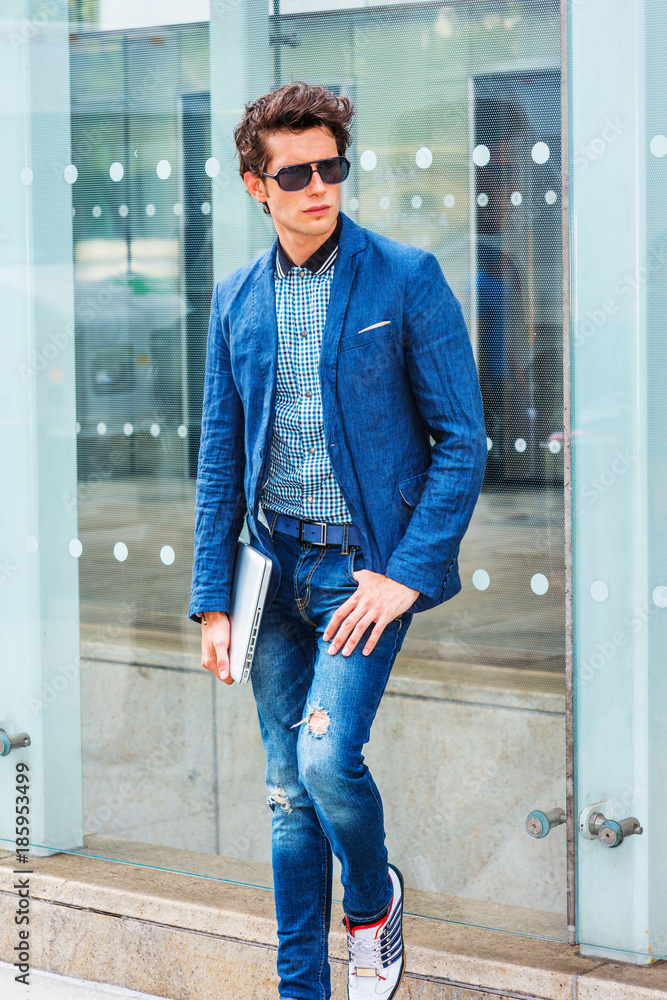European College Student in New Dressing in blue jeans, sneakers, wearing sunglasses, holding laptop computer, a young guy standing against glass wall on street, looking away, thinking.. Stock