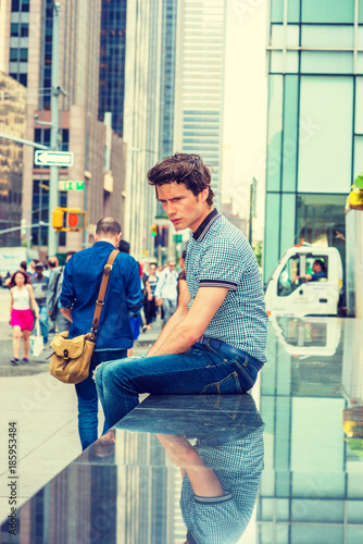 European student traveling in New York. Wearing blue pattered short sleeve shirt, jeans, a guy sitting on street with high buildings, many people, looking down, sad, thinking. Filtered look..