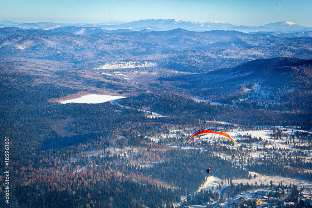 A man flies with an orange parachute, a hang-glider from the top of a snow-covered mountain in a ski resort, from above a beautiful view of the small village of Sheregesh and a ski lift in winter A man flies with an orange parachute, a hang-glider from the top of a snow-covered mountain in a ski resort, from above a beautiful view of the small village of Sheregesh and a ski lift in winter