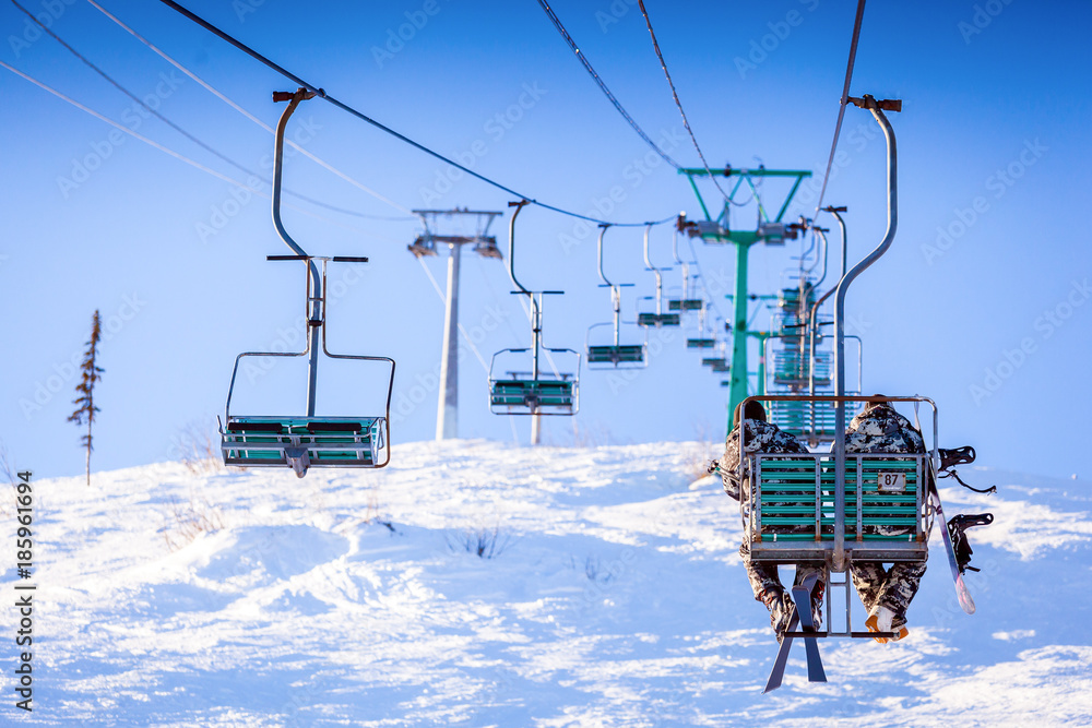 Two men skiers in warm clothes and with mountain skis climb up the ski lift up the mountain in a ski resort on a winter warm day, the back view