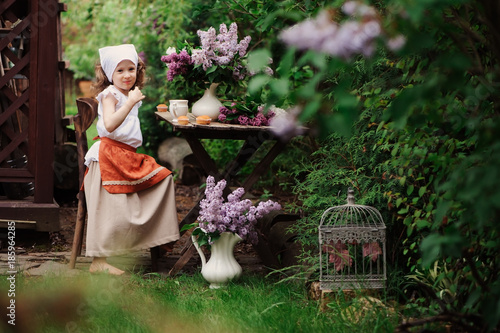 kid girl at garden tea party in spring day with bouquet of lilacs (syringa), rustic wooden table and vintage dress