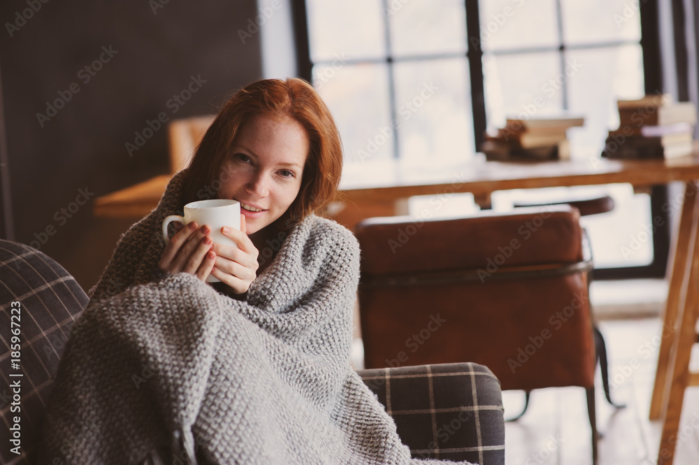 young sick woman healing with hot drink at home on cozy couch, wrapped in knitted blanket - obrazy, fototapety, plakaty 