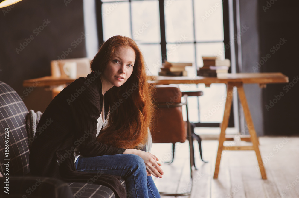 young readhead woman relaxing at home on cozy couch, dressed in casual sweater and jeans. Calm winter or autumn weekend
