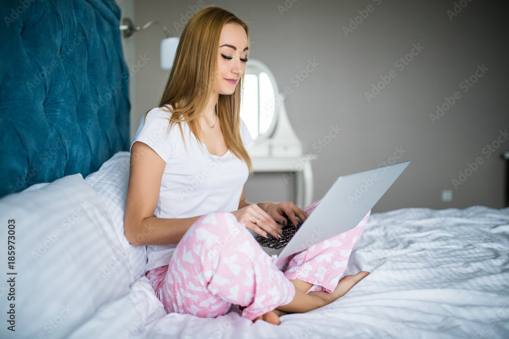 Smiling woman catching up on her social media as she relaxes in bed with a laptop on a lazy day Smiling woman catching up on her social media as she relaxes in bed with a laptop on a lazy day