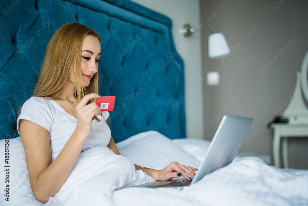 Smiling woman lying on her bed with her credit card in hand as she uses her laptop while wake up in the morning Smiling woman lying on her bed with her credit card in hand as she uses her laptop while wake up in the morning