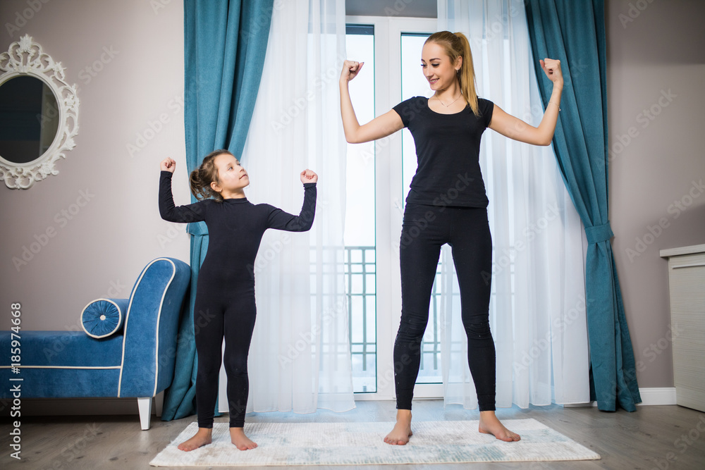 Fototapeta premium Beautiful young woman and charming little daughter are smiling while doing yoga together at home