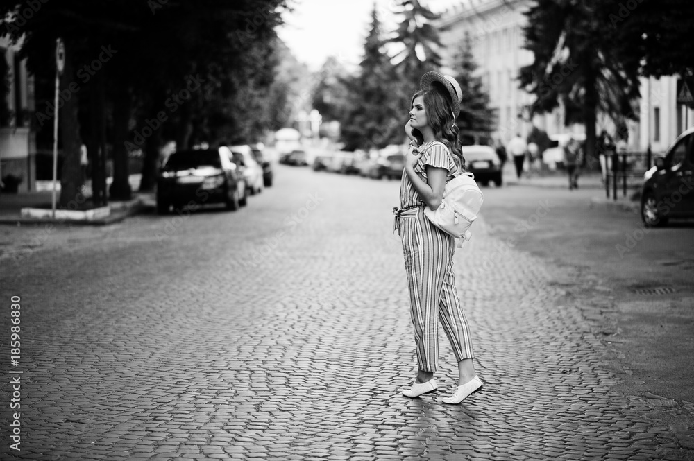 Portrait of a beautiful model in striped overall posing with hat and a backpack on a street with trees in a town. Black and white photo.