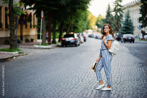 Portrait of a beautiful model in striped overall posing with hat and a backpack on a street with trees in a town. © AS Photo Family