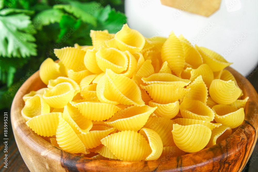 Culinary background with conchiglie pasta on wooden table. Pasta in the form of cockleshells in a wooden bowl with parsley.