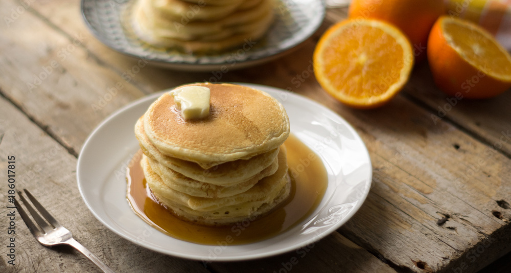 Homemade pancakes with maple syrup and butter on the white plates with oranges on a side, on the wooden table