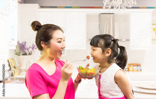 Asian girl in a pink dress is feeding a vegetable salad to a girl in a white kitchen as a background, a healthy diet for girls.