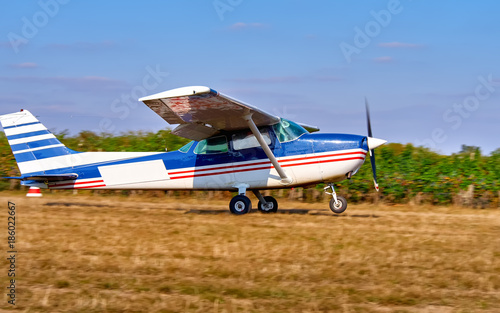 A light aircraft takes off in a field near a vineyard. Sunny summer day, people watch and plant.