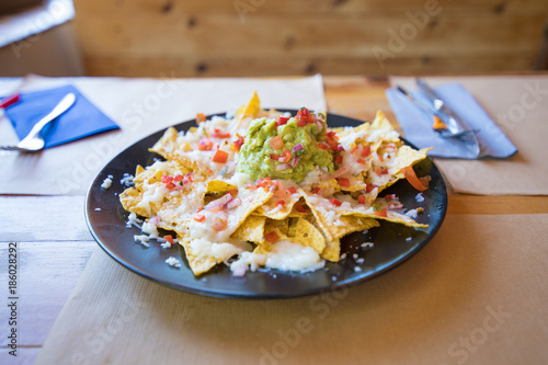 black plate with nachos and guacamole, chopped tomato and cheese, on wooden table with paper placemats at restaurant
