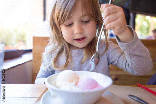 portrait of three years old blonde child ready to eat two scoops of strawberry and vanilla ice cream, in white bowl, with a spoon in raised hand, sitting at the restaurant table
